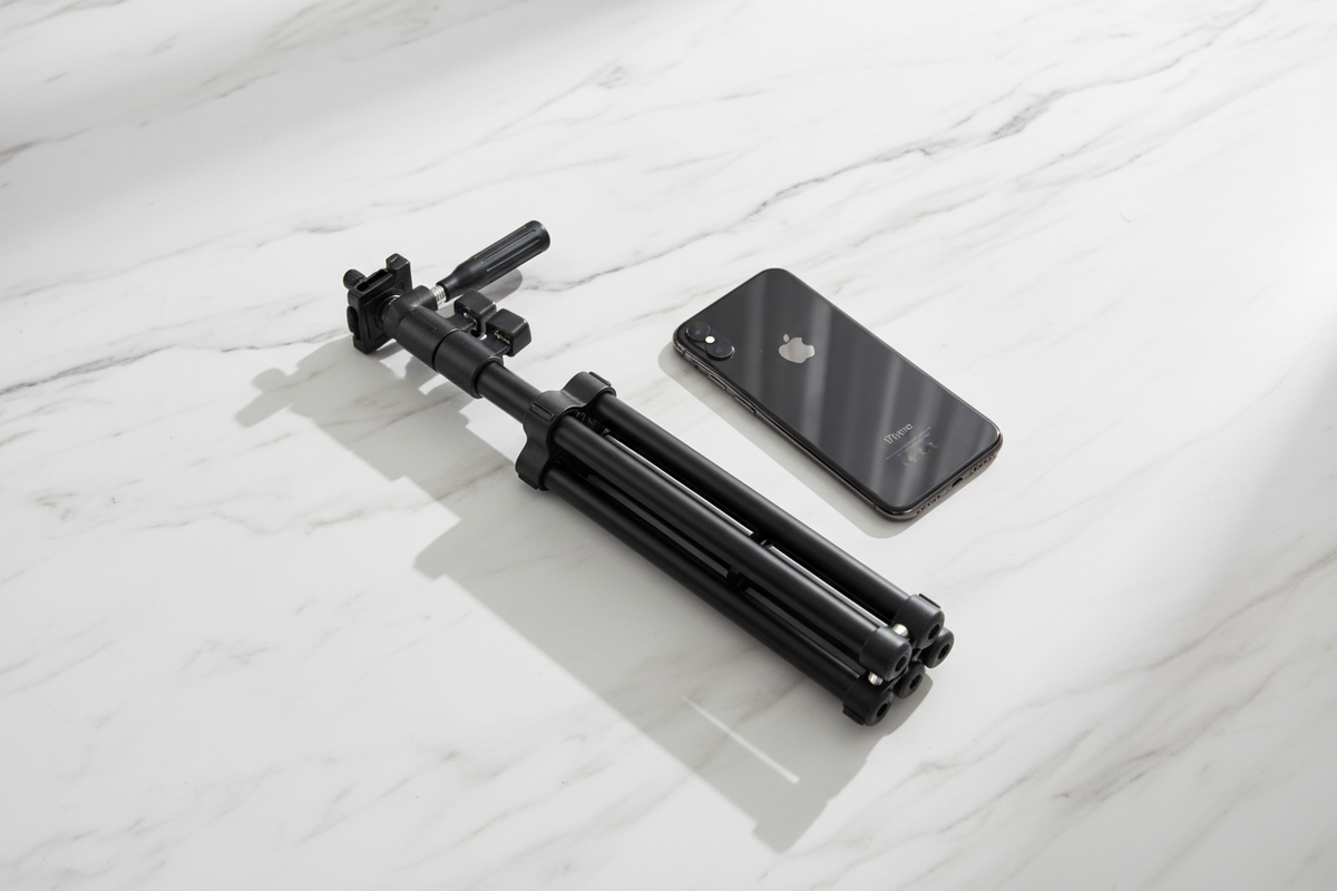 A black smartphone rests next to a collapsed black tripod on a white marble surface.
