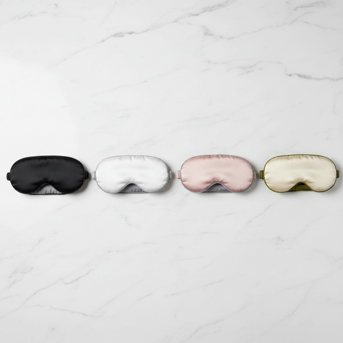 Four silk sleep masks in black, white, pink, and gold lie neatly lined up on a white marble surface.
