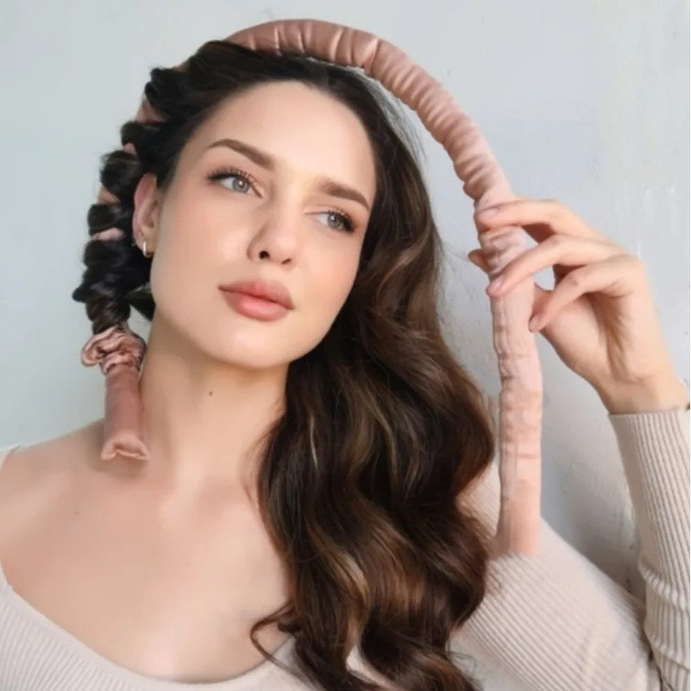 A young woman holds a pink satin heatless hair curler while styling her long brown hair into soft waves.