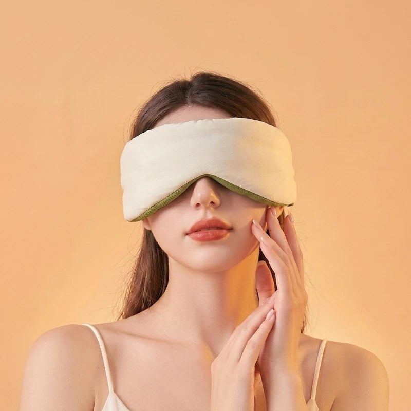 A relaxed woman gently touches her face while wearing a plush, cream-colored weighted sleep mask against a warm peach background.
