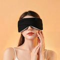 A young woman wears a comfortable black sleep mask and gently touches her cheek against a warm peach background.
