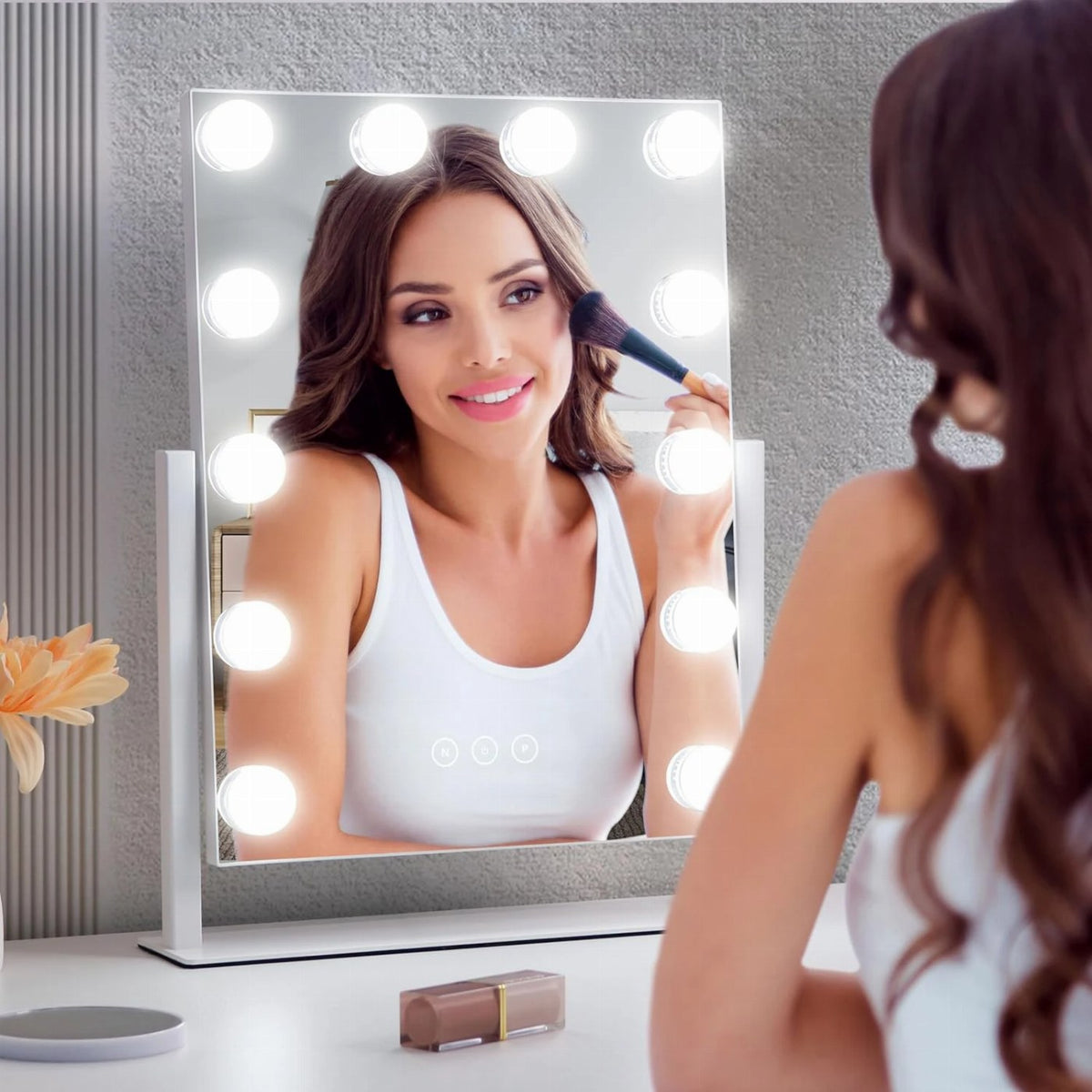 A smiling woman applies makeup using a makeup brush in front of a Hollywood vanity mirror with glowing bulb lights.