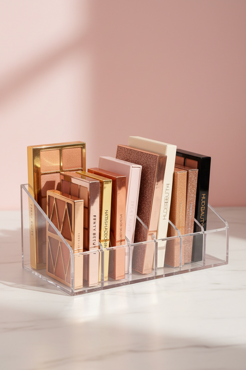 A clear acrylic makeup organizer neatly holds an assortment of colorful eyeshadow palettes on a white marble surface against a soft pink backdrop.